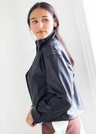 A woman wearing a black faux-leather jacket, photographed from a side-back angle against a bright, neutral indoor background.