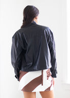 A woman viewed from behind wearing a black leather jacket and a brown and white geometric patterned mini skirt, set against a bright white background.