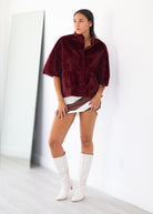 A woman stands in a bright white room wearing a plush burgundy faux-fur cropped jacket, a white and brown mini skirt, and white knee-high boots.