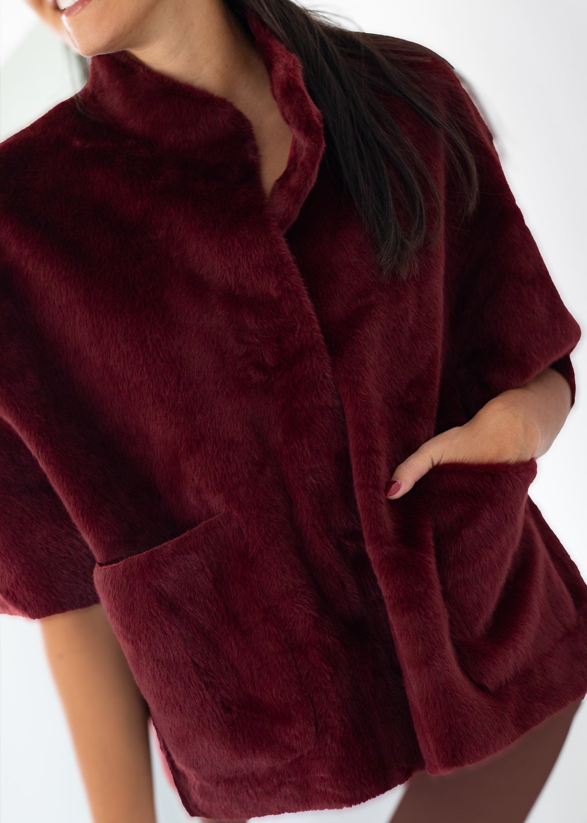 A close-up of a woman wearing a plush, short-sleeved burgundy faux fur jacket with a high collar and front pockets, set against a plain white background.