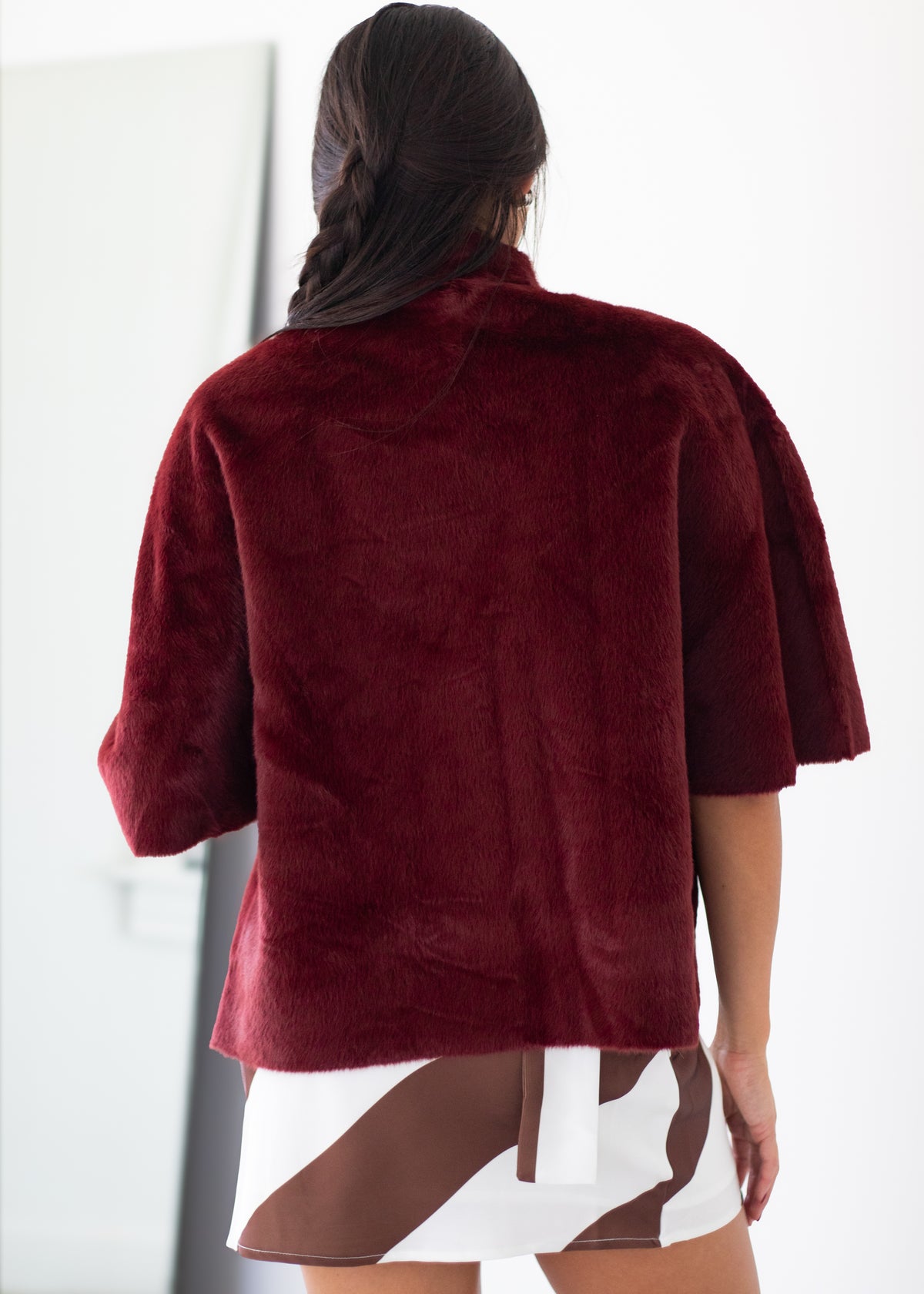 A woman viewed from behind wearing a plush, short-sleeved burgundy faux fur jacket and a brown and white patterned skirt.