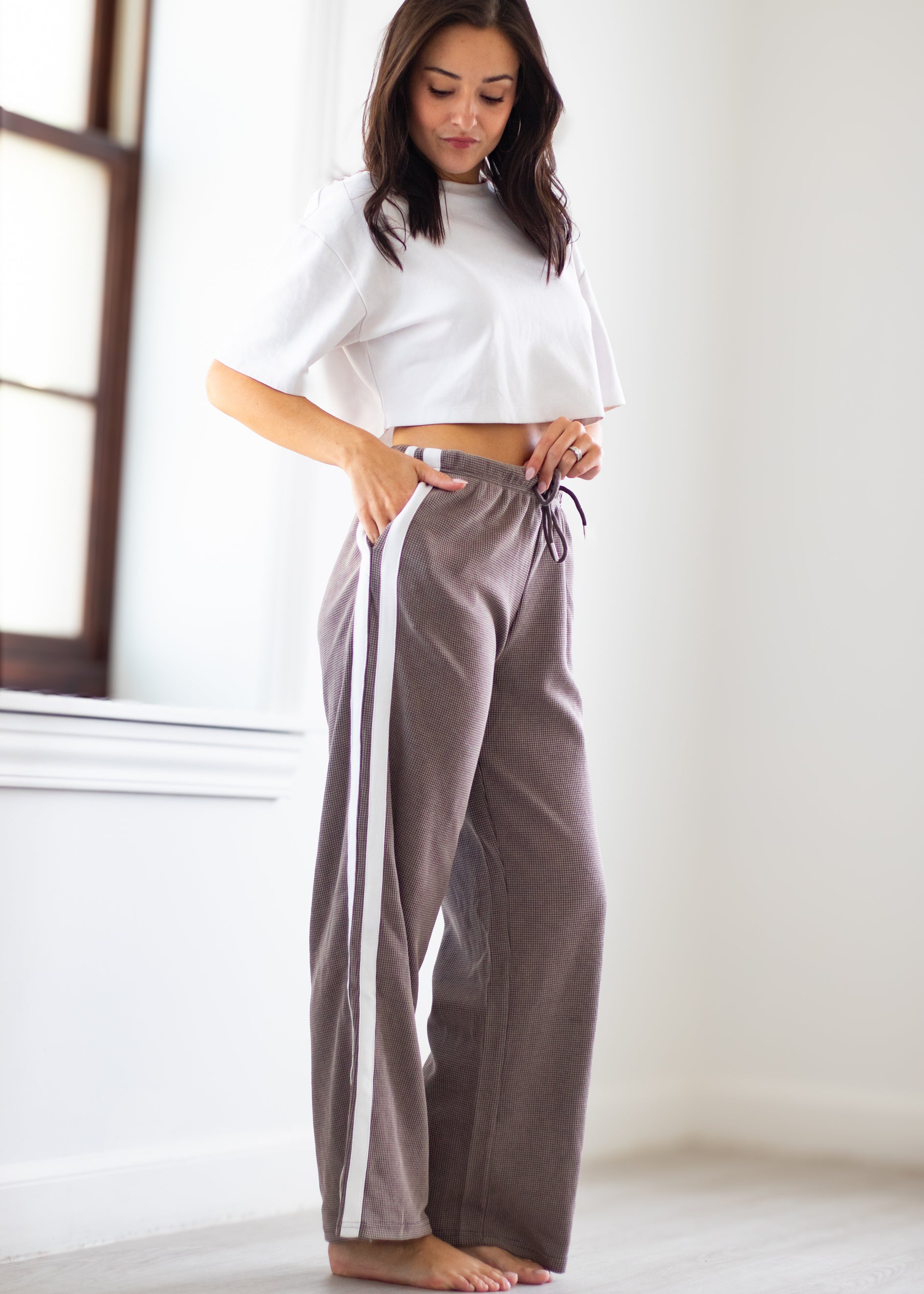 A woman stands barefoot indoors wearing wide-leg taupe pants with white side stripes and a white cropped short-sleeve top.