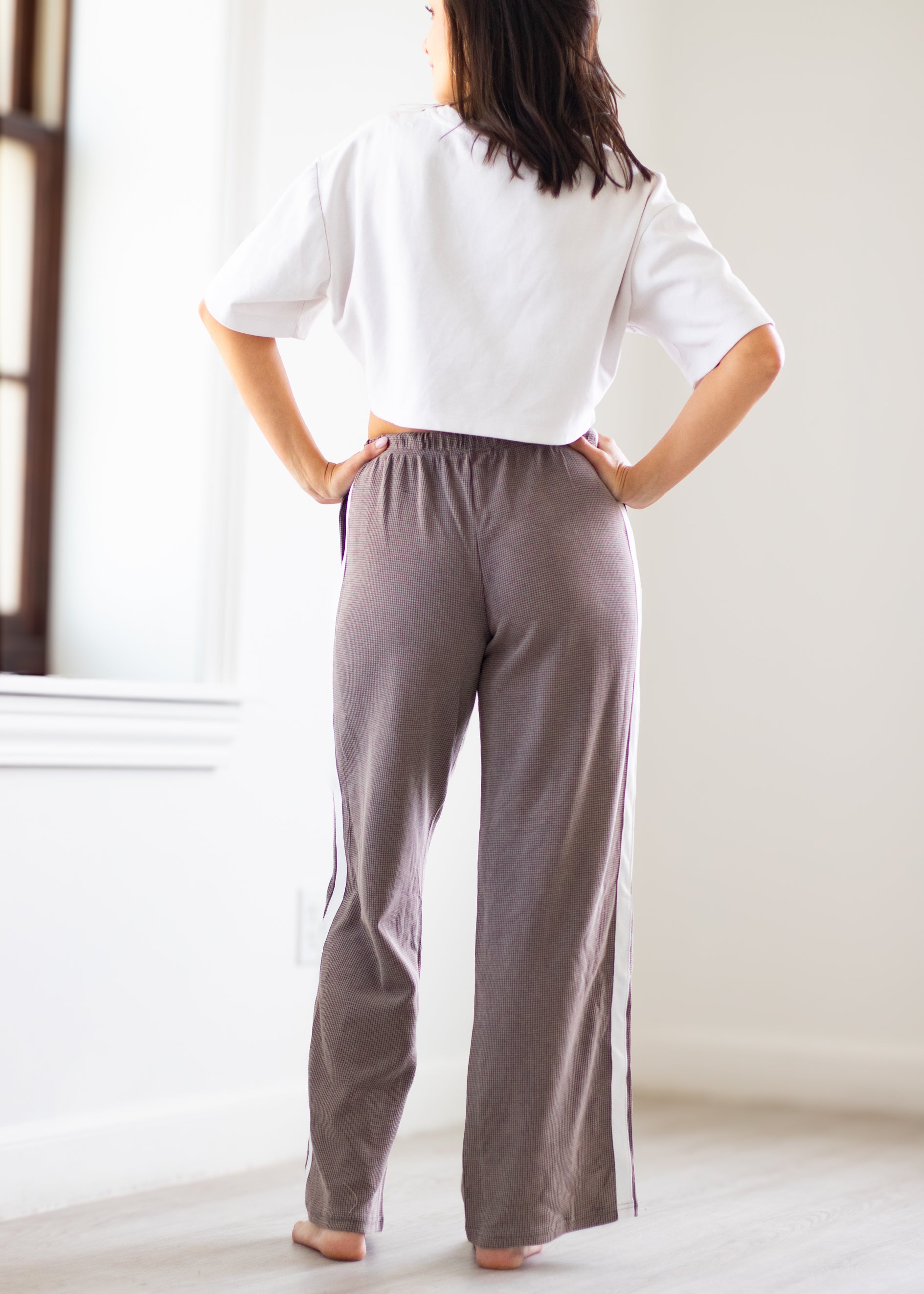 A woman stands with her back to the camera in a bright room, wearing loose, wide-leg lounge pants in a muted taupe-gray color with white side stripes, paired with a white cropped t-shirt.