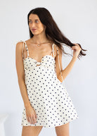 A woman modeling a white mini dress with black polka dots and thin tie-shoulder straps against a plain light gray studio background.