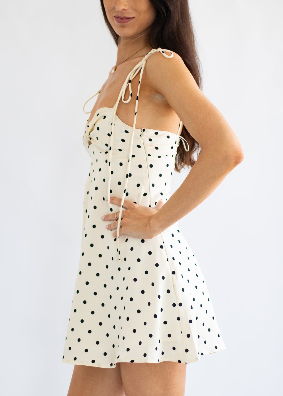 A woman wearing a white sleeveless mini dress with black polka dots and thin adjustable tie straps, shown against a plain light background.