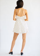 A woman viewed from behind wearing a white spaghetti-strap mini dress with black polka dots and black block-heel mules, standing against a plain white studio background.
