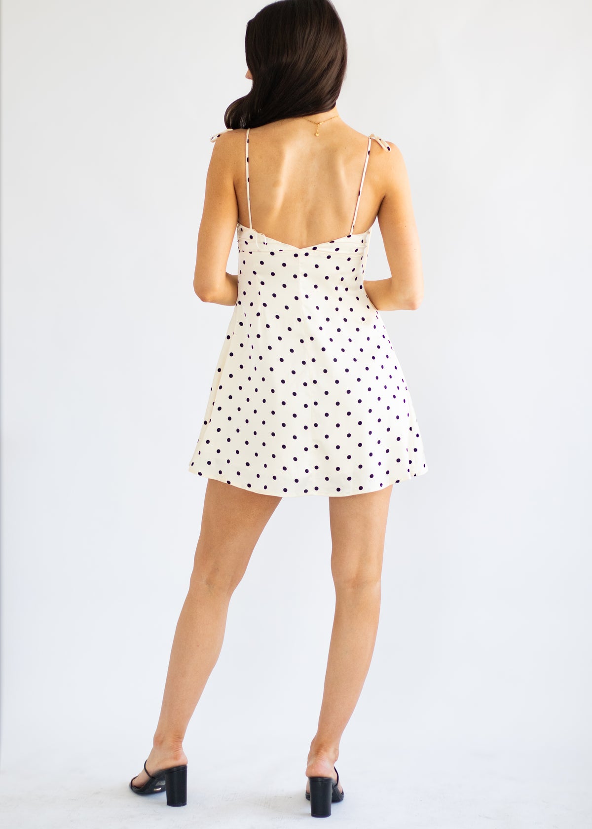 A woman viewed from behind wearing a white spaghetti-strap mini dress with black polka dots and black block-heel mules, standing against a plain white studio background.