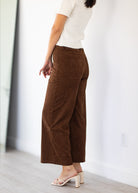 A person shown from the back wearing brown wide-leg cropped pants, a white short-sleeve top, and light pink heeled mule sandals in a bright indoor room with a white wall and light wood floor.