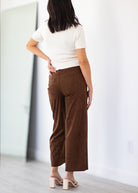 A woman shown from behind wearing brown wide-leg cropped pants, a white short-sleeve top, and beige block-heel mules in a bright indoor room.