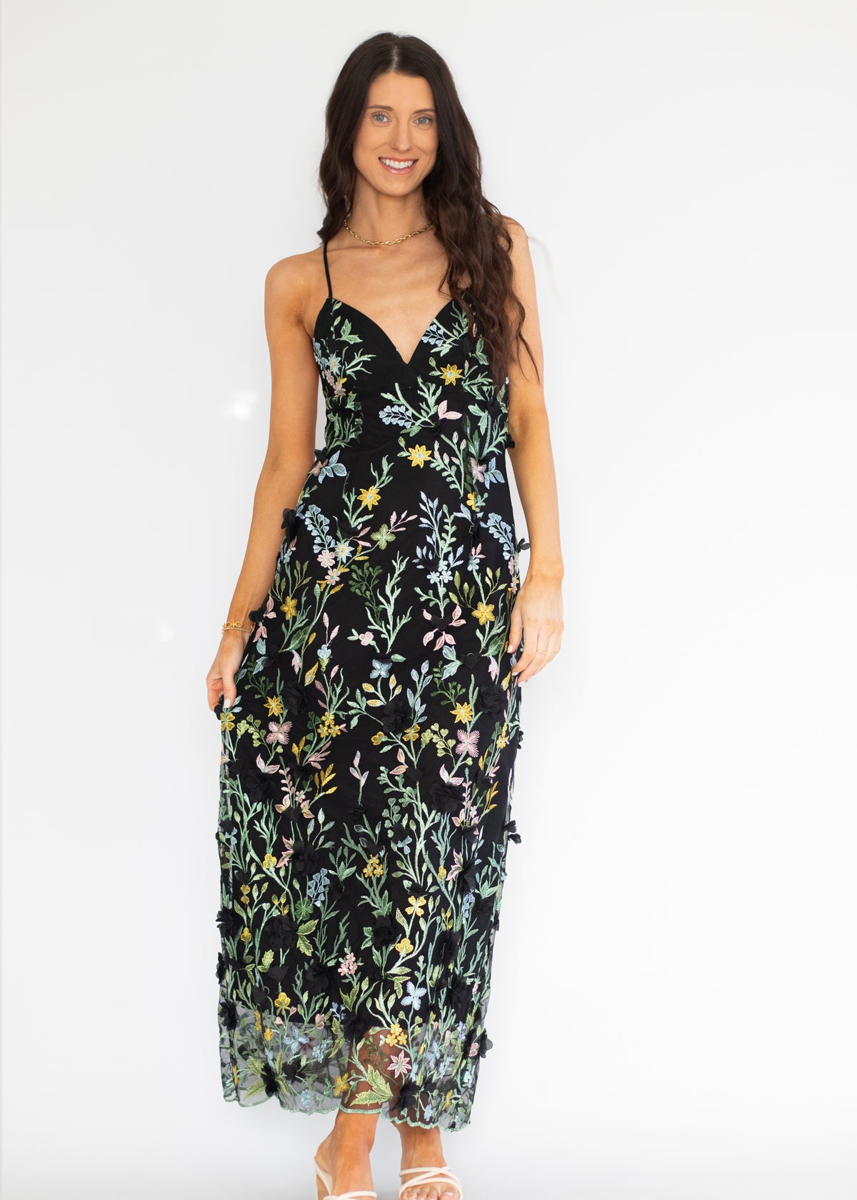 A woman modeling a black midi slip dress with a colorful floral and butterfly print, featuring thin spaghetti straps and a V-neckline, set against a plain white studio background.