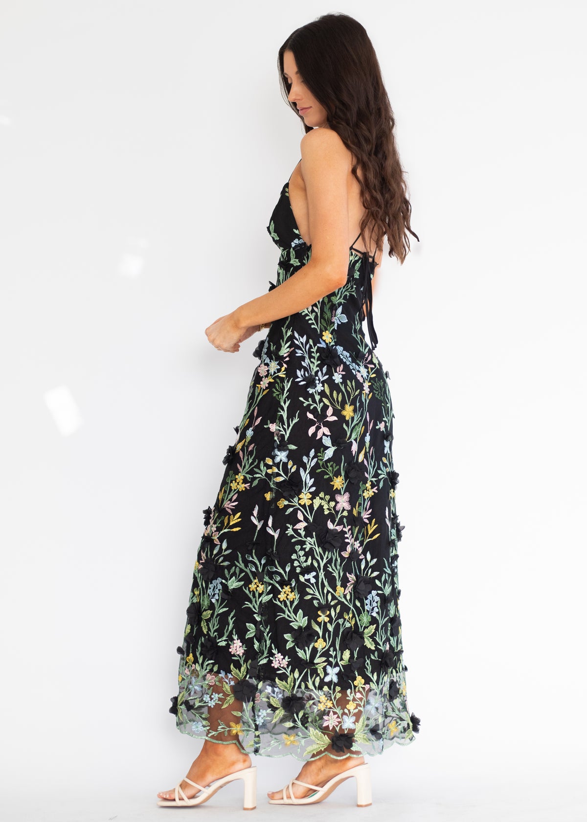 A woman modeling a sleeveless black maxi dress with a colorful floral print and ruffled tulle hem, paired with white strappy high-heeled sandals against a plain white background.