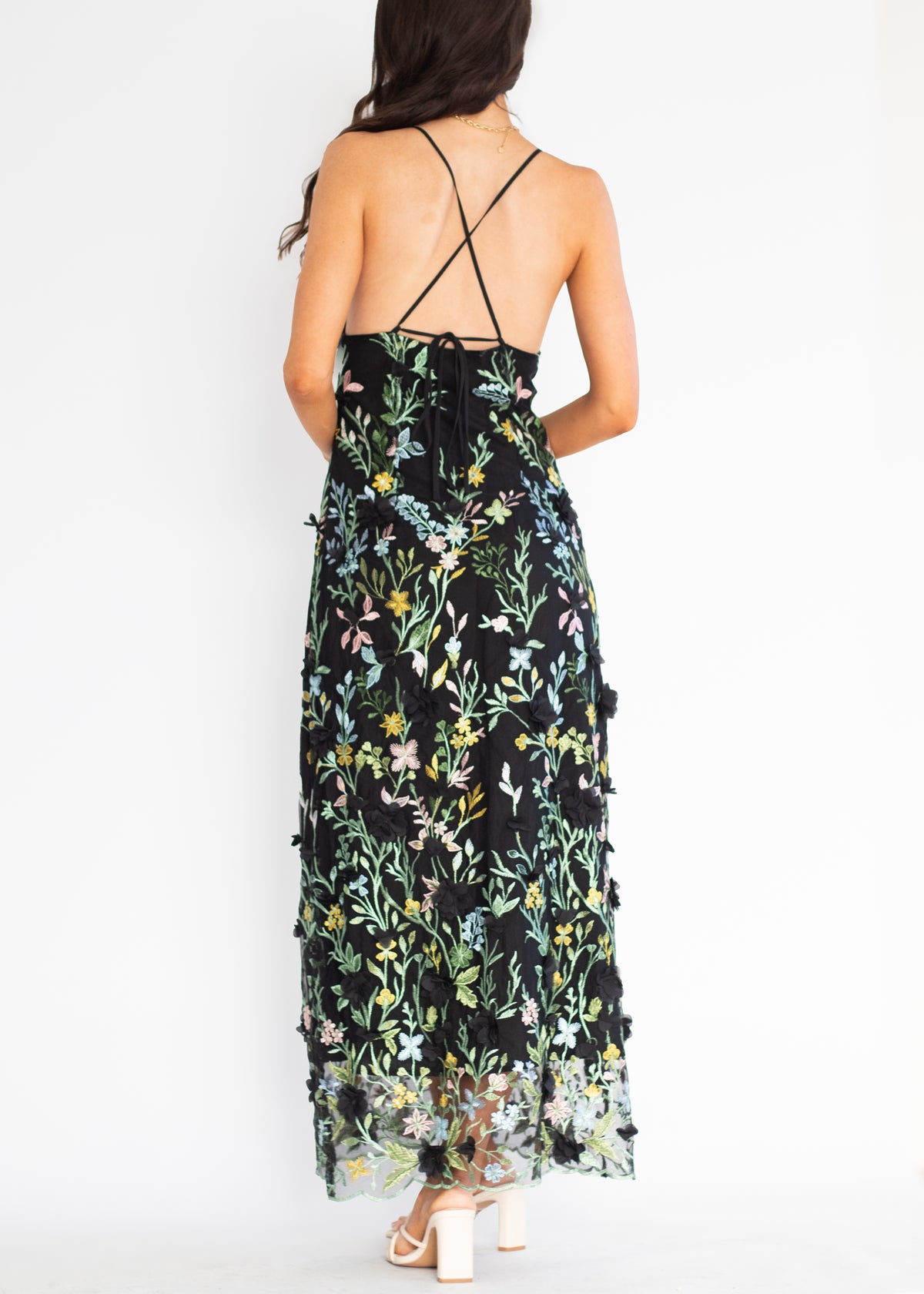 A woman shown from the back wearing a sleeveless black maxi dress with a colorful floral print and thin crisscross back straps, paired with white block-heel sandals against a plain white studio background.