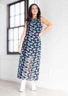 A woman modeling a sleeveless navy maxi dress with a light blue and brown floral pattern, paired with white ankle boots, standing in a bright room with a large window.