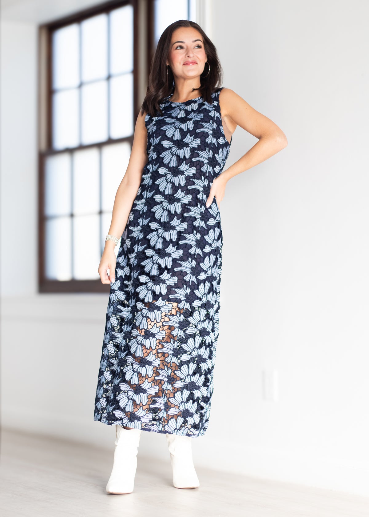 A woman modeling a sleeveless navy maxi dress with a light blue and brown floral pattern, paired with white ankle boots, standing in a bright room with a large window.