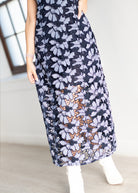 A woman wearing a sleeveless navy maxi skirt with a white floral pattern and a sheer lace overlay near the hem, paired with white ankle boots.