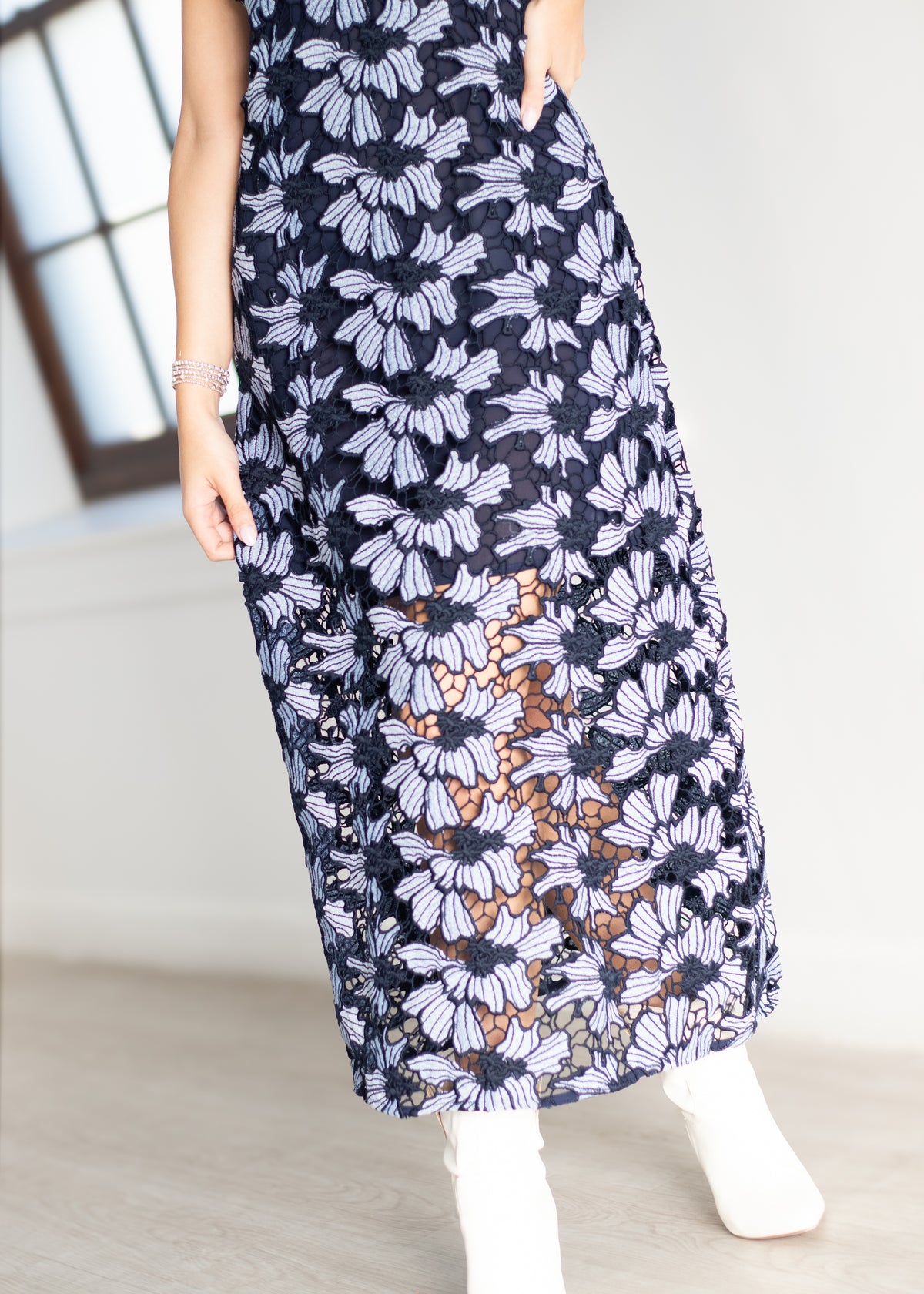 A woman wearing a sleeveless navy maxi skirt with a white floral pattern and a sheer lace overlay near the hem, paired with white ankle boots.