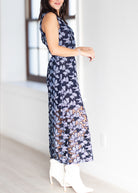 A woman modeling a sleeveless navy maxi dress with a purple and orange floral lace overlay, paired with white heeled boots in a bright indoor setting.