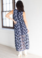 A woman viewed from behind wearing a sleeveless navy maxi dress with a white floral pattern, standing in a bright room with a window and light-colored floor.