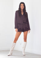 A woman modeling a dark plum purple and white polka dot long-sleeve romper with lace trim, paired with white knee-high heeled boots, in a bright white room.
