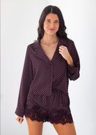 A woman modeling a dark plum purple and white polka dot long-sleeve shirt with matching lace-trimmed shorts against a plain white background.