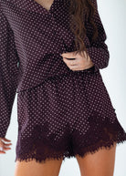 A close-up of a model wearing a matching dark plum pajama set with white polka dots, featuring a long-sleeve top and shorts with lace trim.
