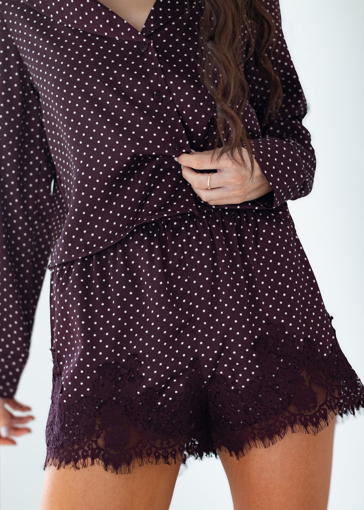 A close-up of a model wearing a matching dark plum pajama set with white polka dots, featuring a long-sleeve top and shorts with lace trim.