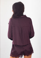 A woman shown from the back wearing a matching long-sleeve button-up shirt and shorts set in a deep purple color with small white polka dots and a lace-trimmed hem, against a plain white background.
