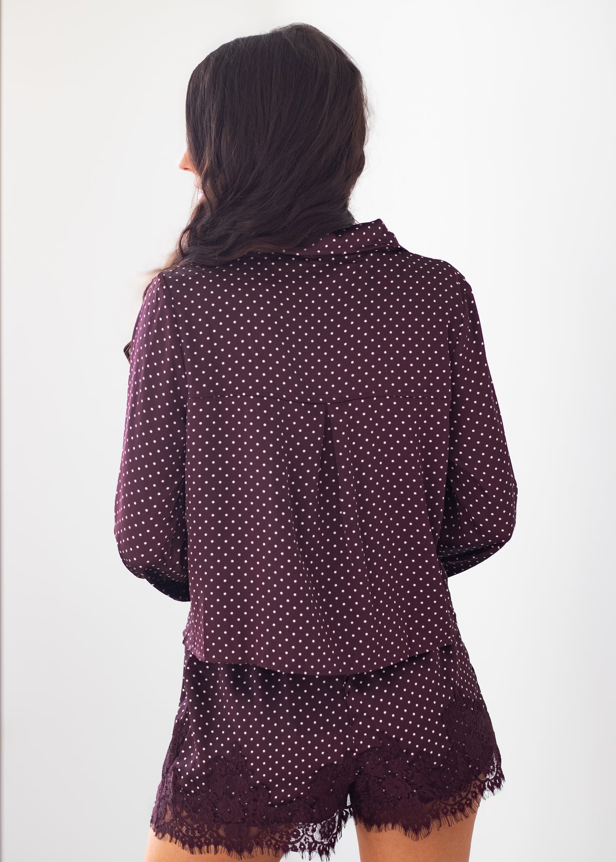 A woman shown from the back wearing a matching long-sleeve button-up shirt and shorts set in a deep purple color with small white polka dots and a lace-trimmed hem, against a plain white background.
