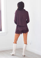 A woman shown from behind wearing a matching dark purple and white polka dot long-sleeve top and shorts set, paired with white knee-high boots, in a bright room with white walls and light wood flooring.