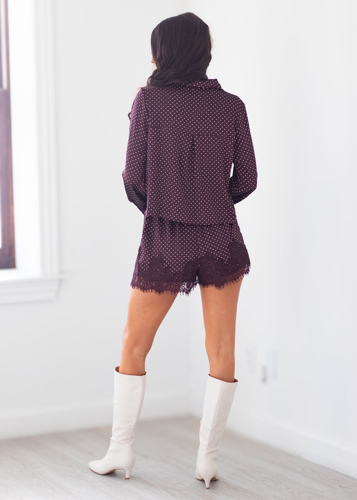 A woman shown from behind wearing a matching dark purple and white polka dot long-sleeve top and shorts set, paired with white knee-high boots, in a bright room with white walls and light wood flooring.
