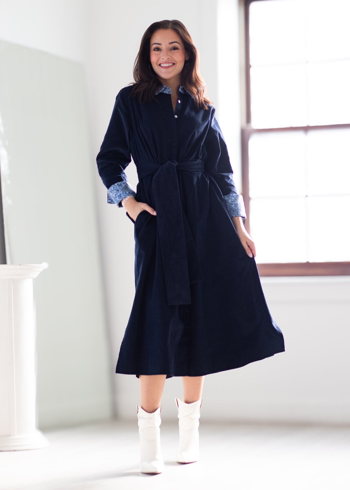 A woman wearing a dark navy blue velvet robe dress with a tie waist and button collar, layered over a blue patterned shirt, paired with white ankle boots, standing in a bright room with a window.