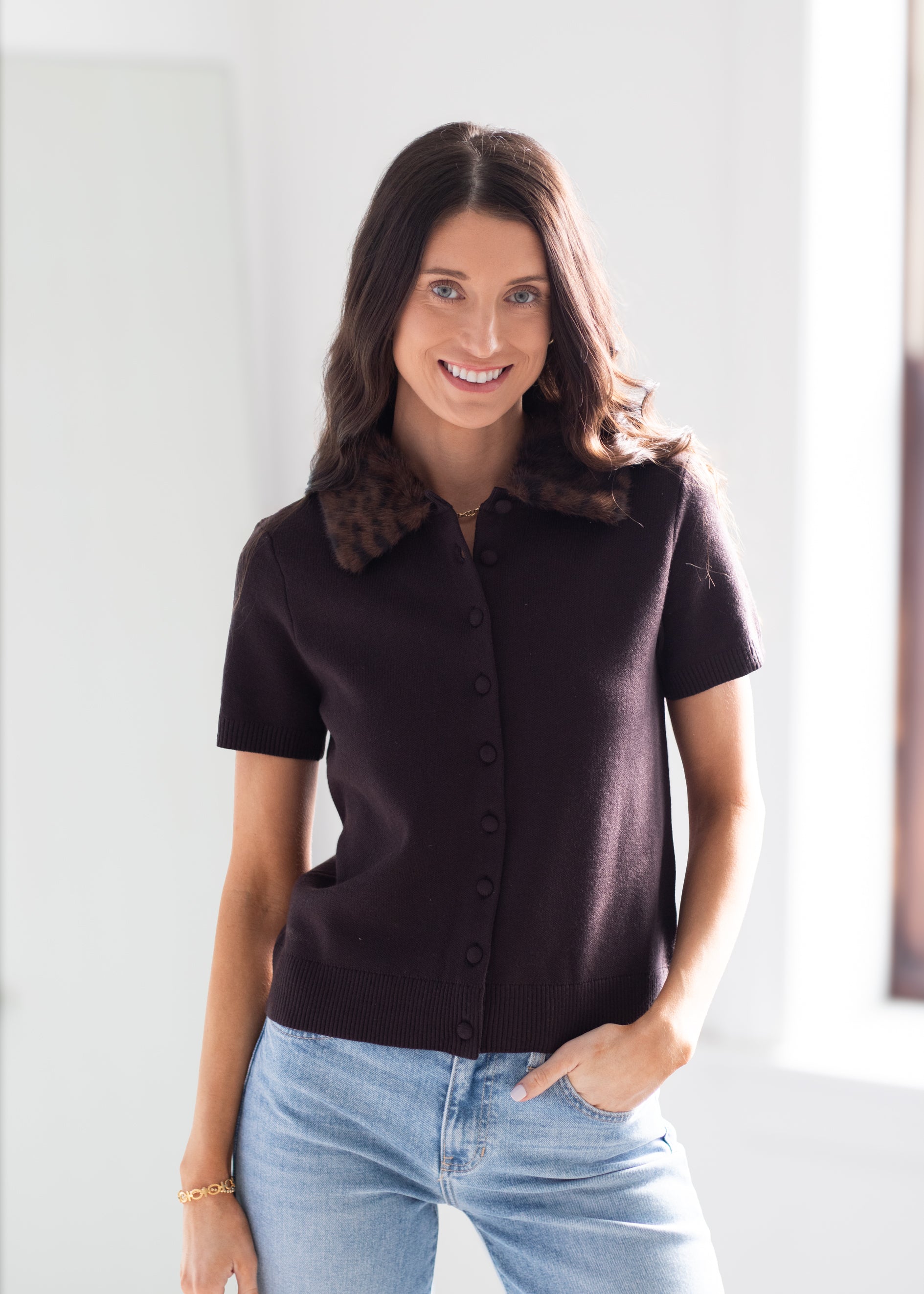 A woman wearing a short-sleeve dark brown knit polo shirt with a brown patterned collar, paired with light blue jeans, standing in a brightly lit indoor setting.