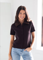 A woman wearing a short-sleeve dark brown knit polo shirt with a brown patterned collar, paired with light blue jeans, standing in a brightly lit indoor setting.