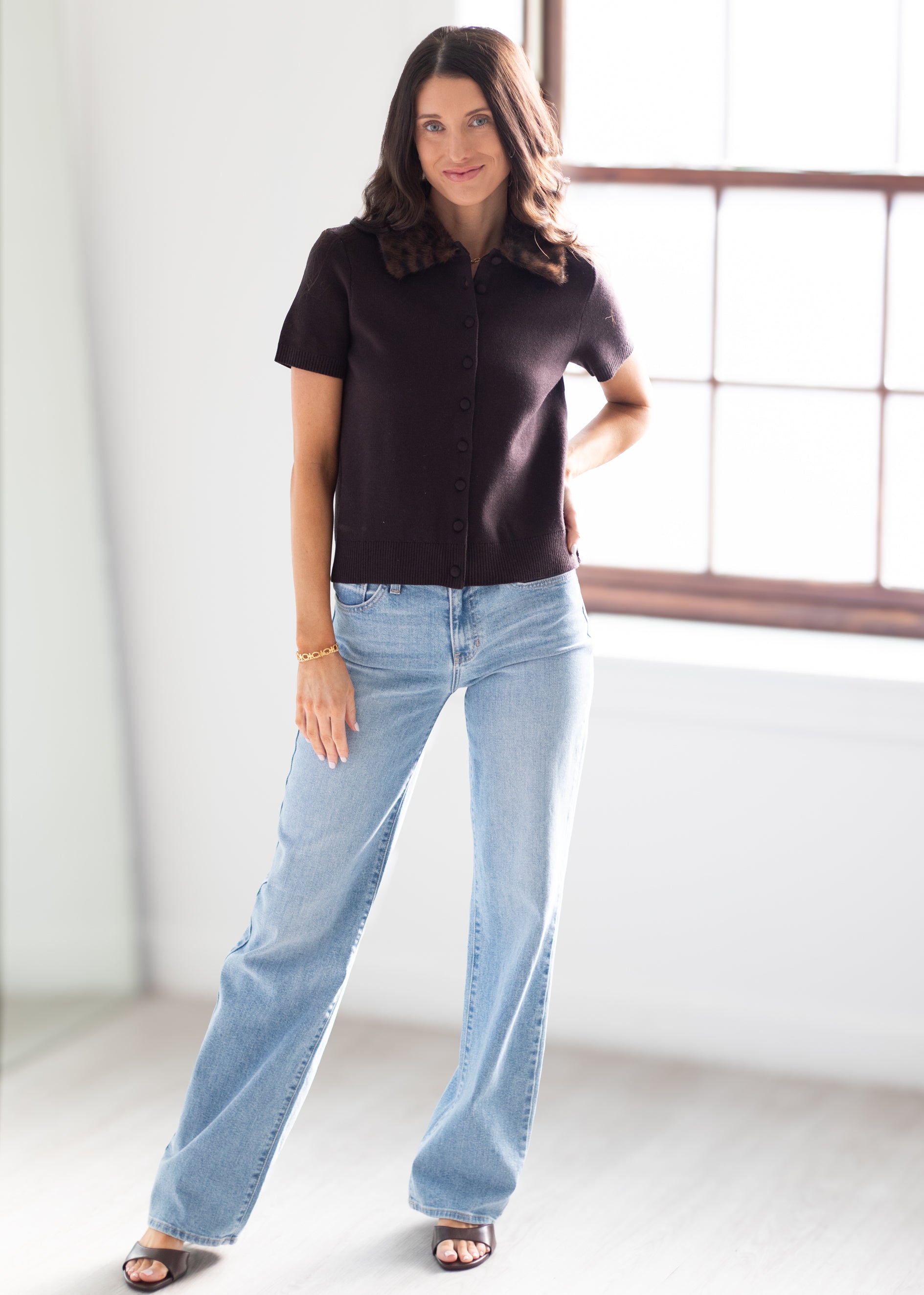A woman modeling a short-sleeve dark brown button-up shirt and light-wash blue jeans, standing in a bright room with a large window.