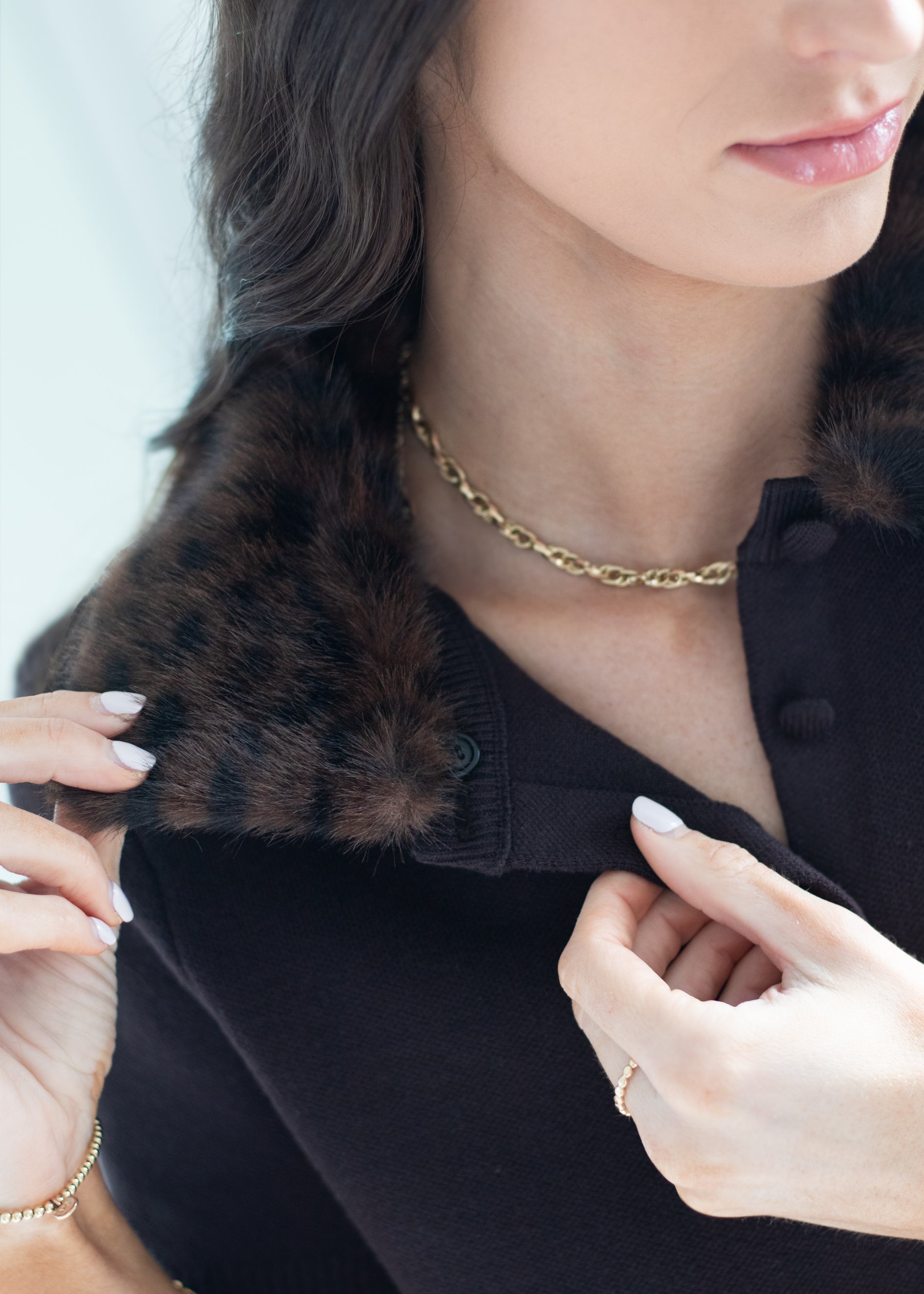 Close-up of a woman wearing a black knit top with a dark brown fur collar, accessorized with a gold chain necklace and bracelet.