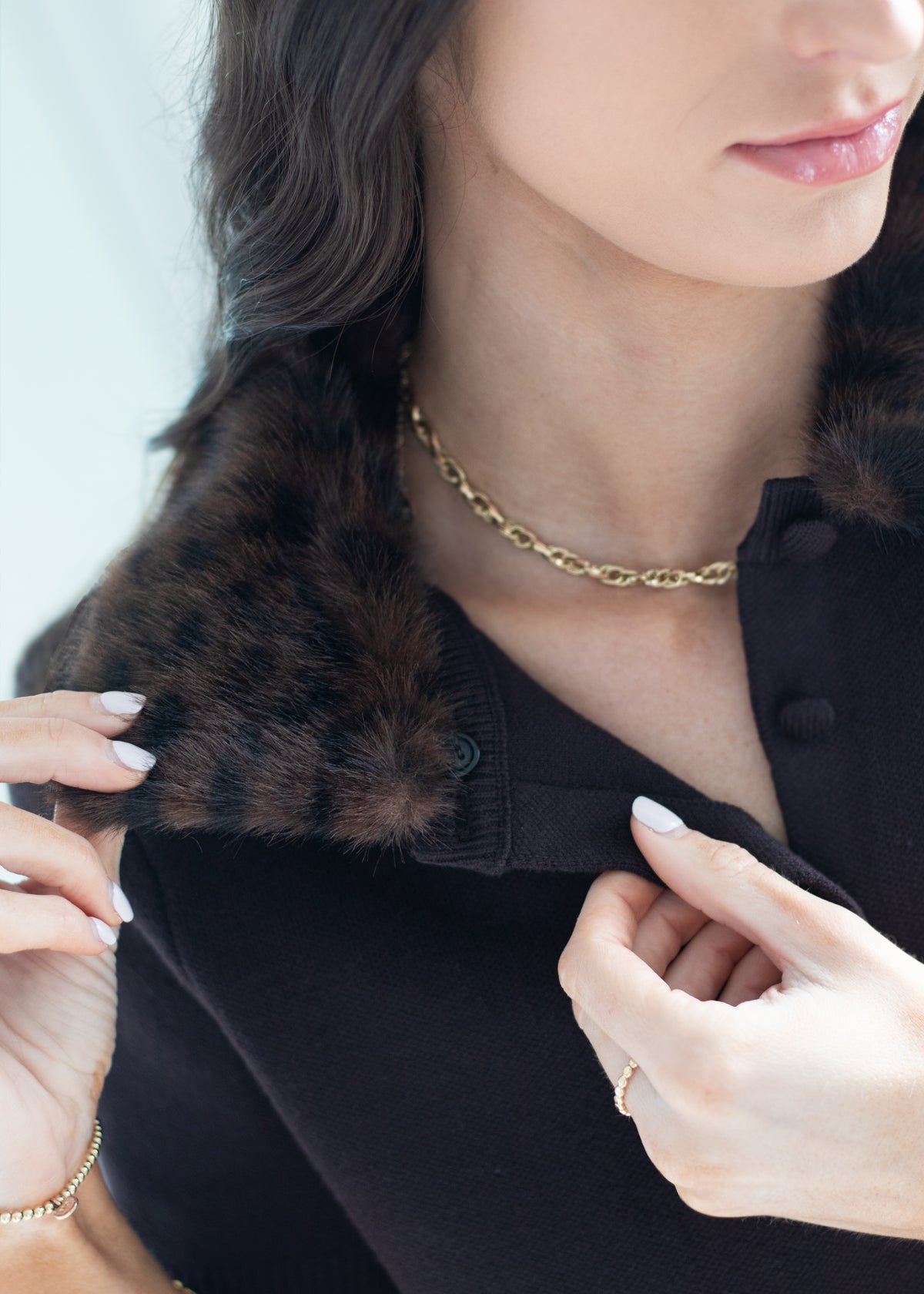 Close-up of a woman wearing a black knit top with a dark brown fur collar, accessorized with a gold chain necklace and bracelet.