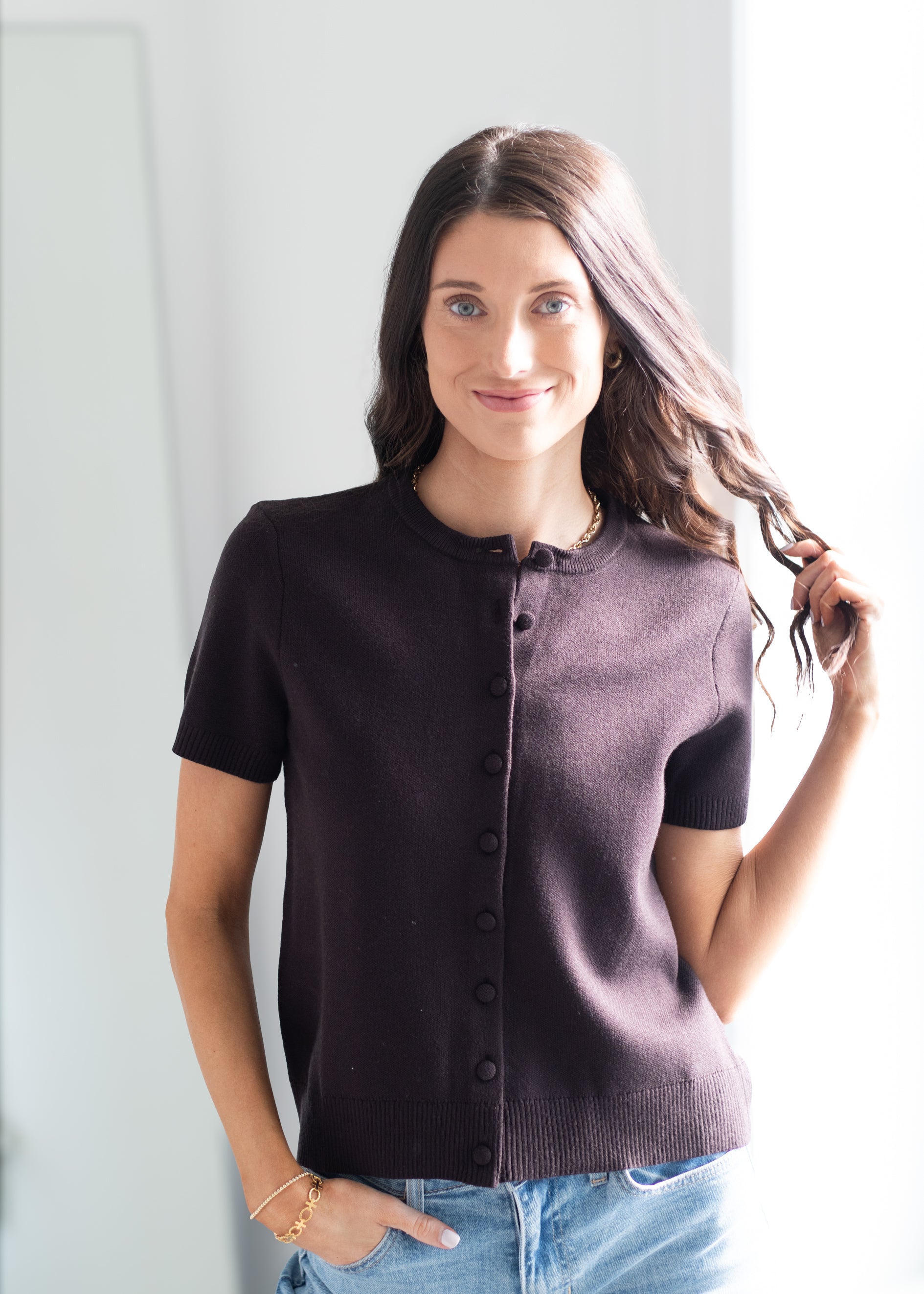 A woman modeling a short-sleeve button-front knit top in a very dark plum or eggplant shade, paired with light blue jeans, standing against a bright white background.