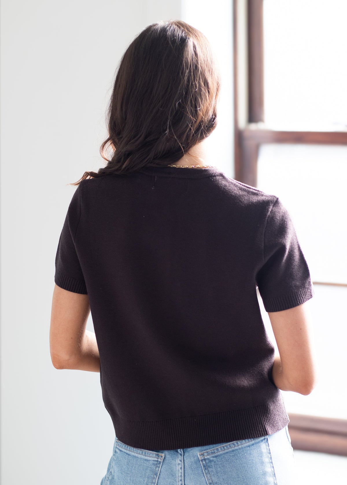 A woman viewed from behind wearing a short-sleeve knit top in a very dark plum or eggplant shade, paired with light blue jeans, standing near a window.