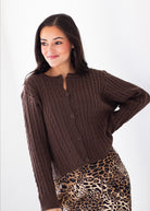 A woman wearing a dark brown cable-knit cropped cardigan with a buttoned neckline, paired with a leopard print skirt, against a plain white studio background.