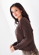 A woman wearing a brown cable-knit long-sleeve sweater and a leopard print skirt, posing against a plain white background.