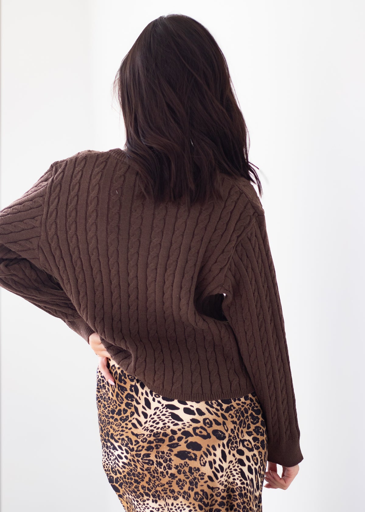 A woman shown from behind wearing a dark brown cable-knit cropped sweater and a leopard-print skirt against a plain white background.