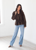 A woman modeling a dark brown belted wrap jacket with voluminous sleeves, paired with light blue wide-leg jeans and black sandals, standing in a bright, minimalist room.