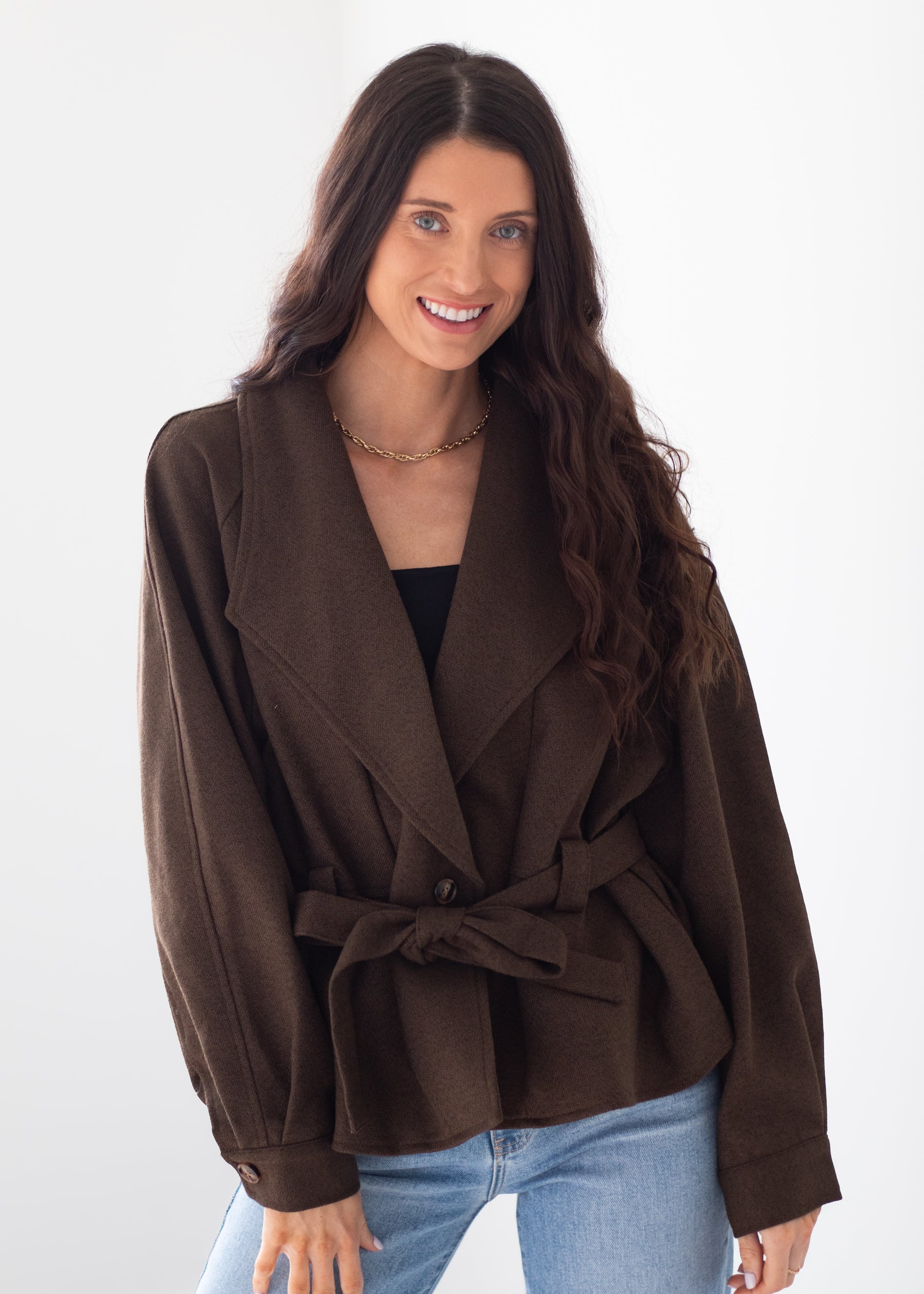 A woman modeling a dark brown belted wrap jacket with wide lapels, worn over a black top and light blue jeans, against a plain white studio background.