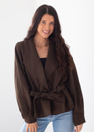 A woman modeling a dark brown belted wrap jacket with wide lapels, worn over a black top and light blue jeans, against a plain white studio background.