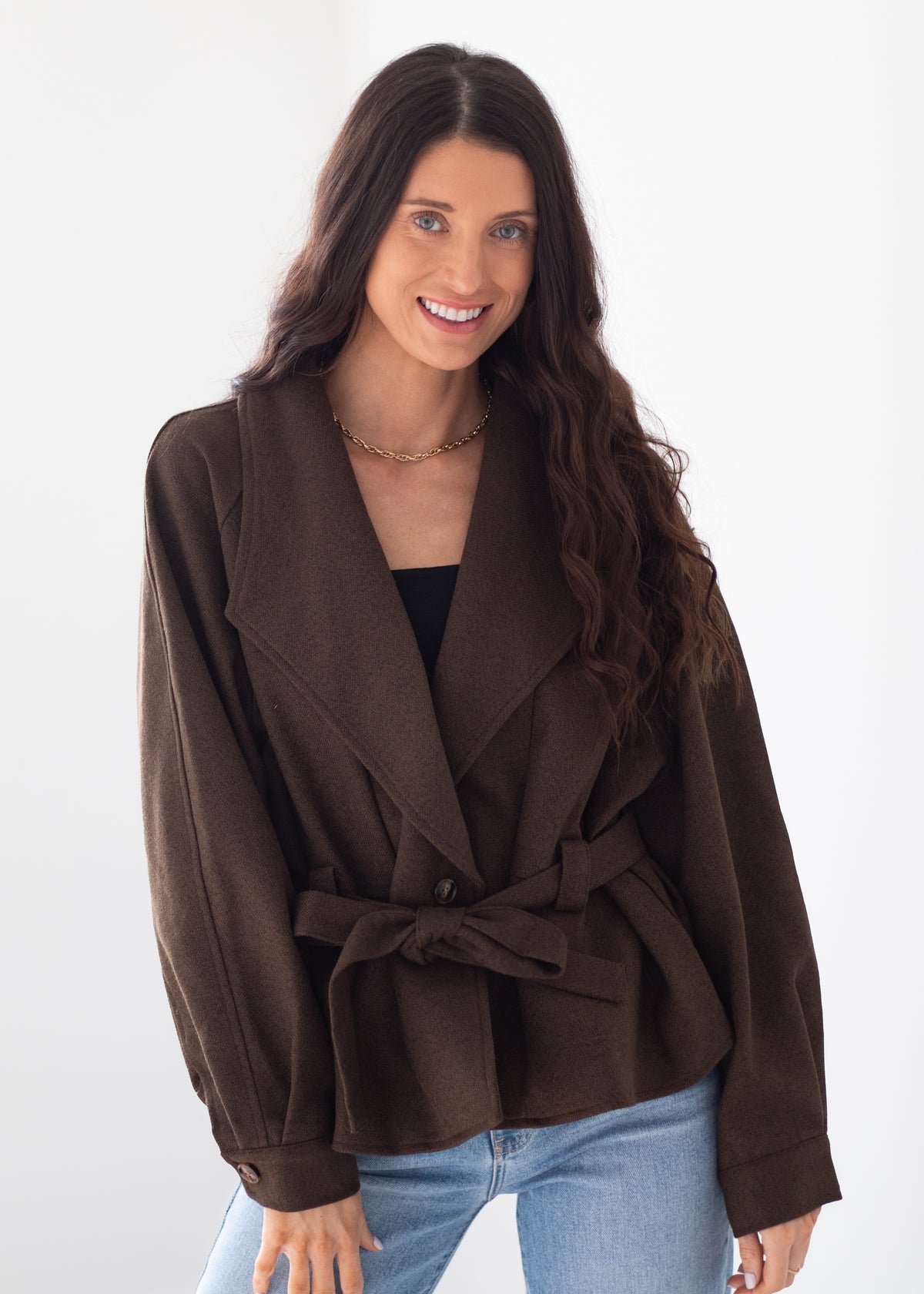 A woman modeling a dark brown belted wrap jacket with wide lapels, worn over a black top and light blue jeans, against a plain white studio background.