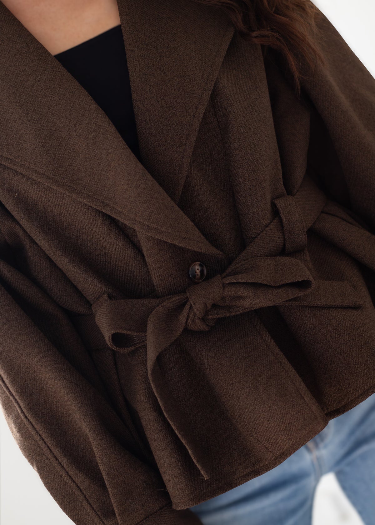 A close-up of a person wearing a dark brown belted wrap jacket over a black top and blue jeans, focusing on the fabric texture and waist tie detail.