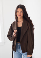 A woman modeling a dark brown open-front wrap jacket over a black top and light blue jeans, standing against a bright white background.