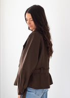 A woman with long dark wavy hair is shown from the side/back wearing a brown belted blazer over light blue jeans, photographed against a plain light background.