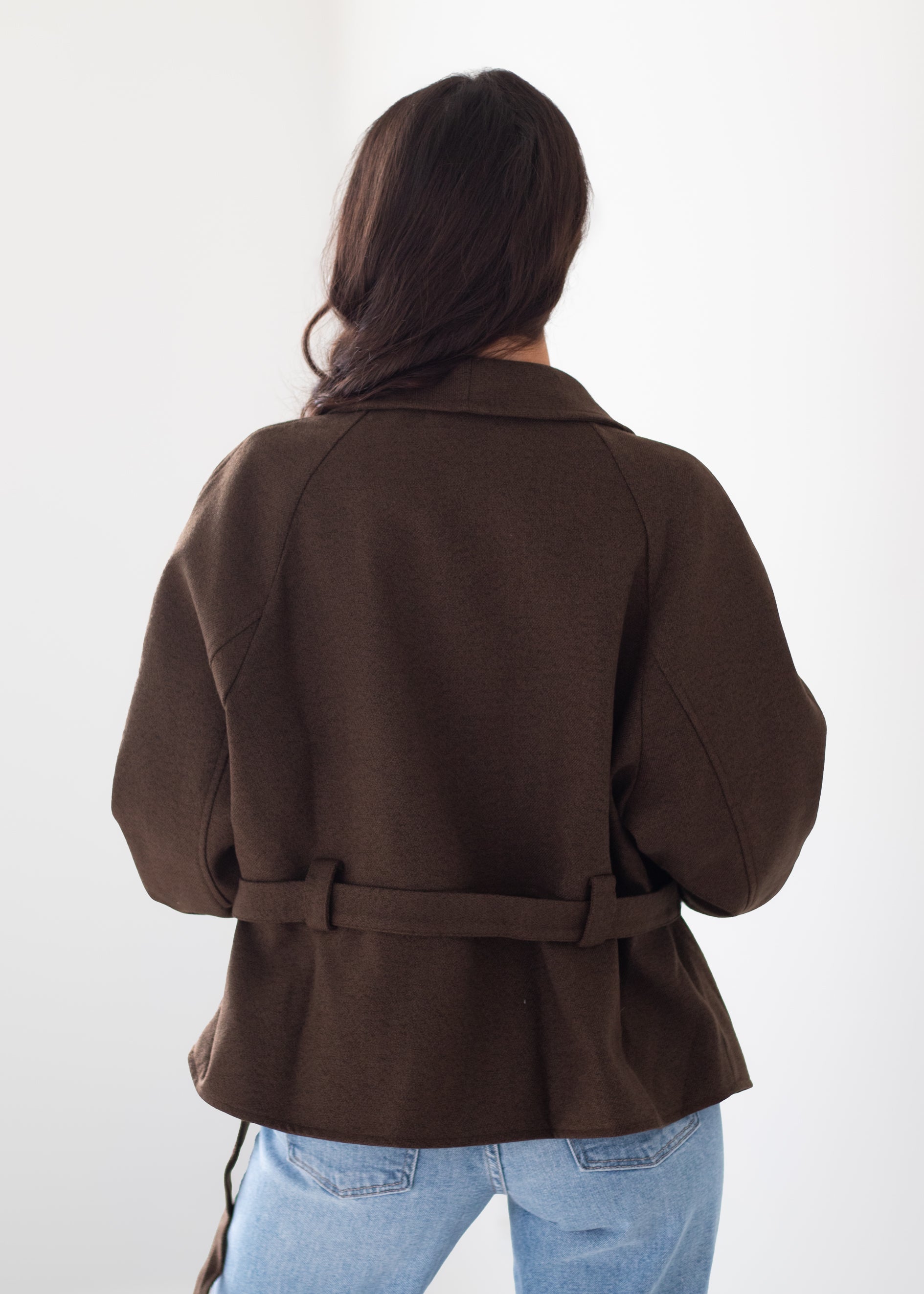 A woman viewed from behind wearing a dark brown belted jacket and light blue jeans against a plain white background.