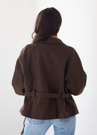 A woman viewed from behind wearing a dark brown belted jacket and light blue jeans against a plain white background.